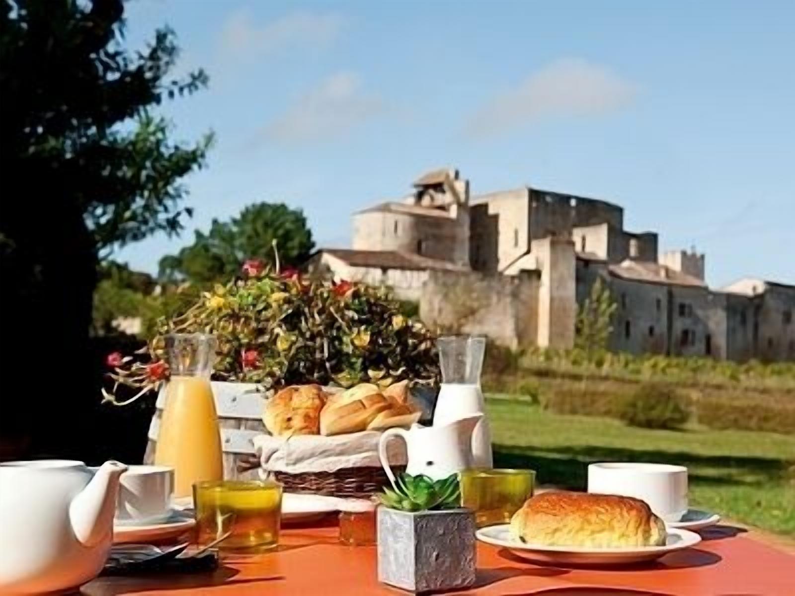 Mobiliario de comedor al aire libre con una mesa, tetera, tazas y una cesta de pan, con un edificio histórico de fondo.