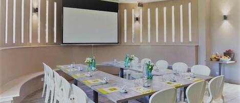 Un salón de conferencias con una larga mesa, sillas blancas, una pantalla de proyector e iluminación empotrada.