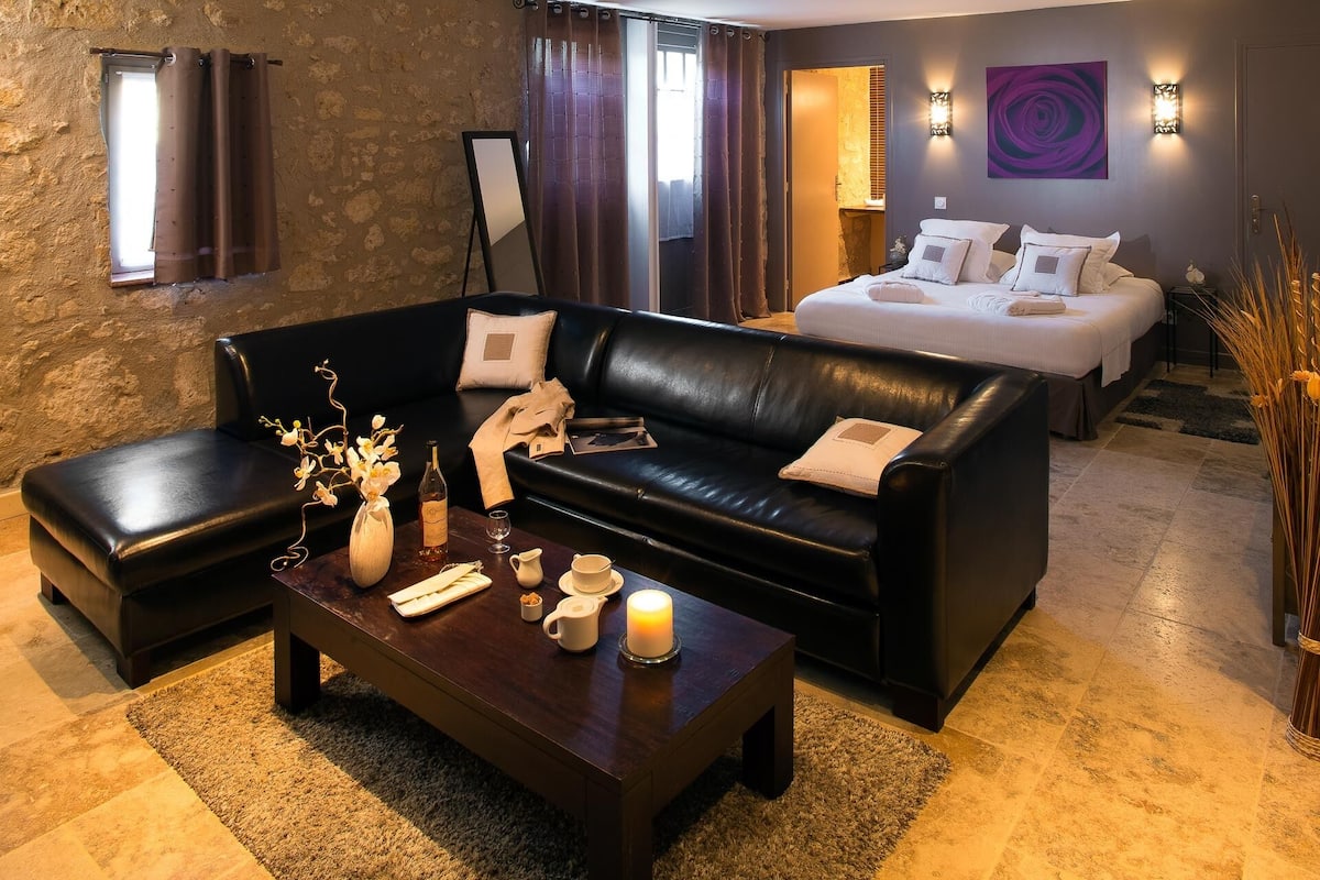 A modern hotel room with a black leather sofa, a wooden coffee table with decorative items, a bed with white bedding, and a stone wall.