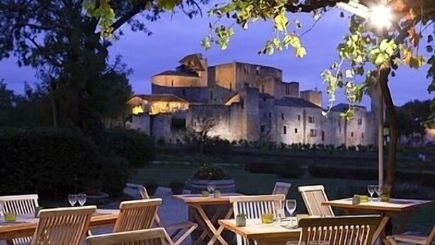 An outdoor dining area with tables and chairs, a view of a castle-like building in the background, and a tree overhead. - L'Auberge de Larressingle (Larressingle)