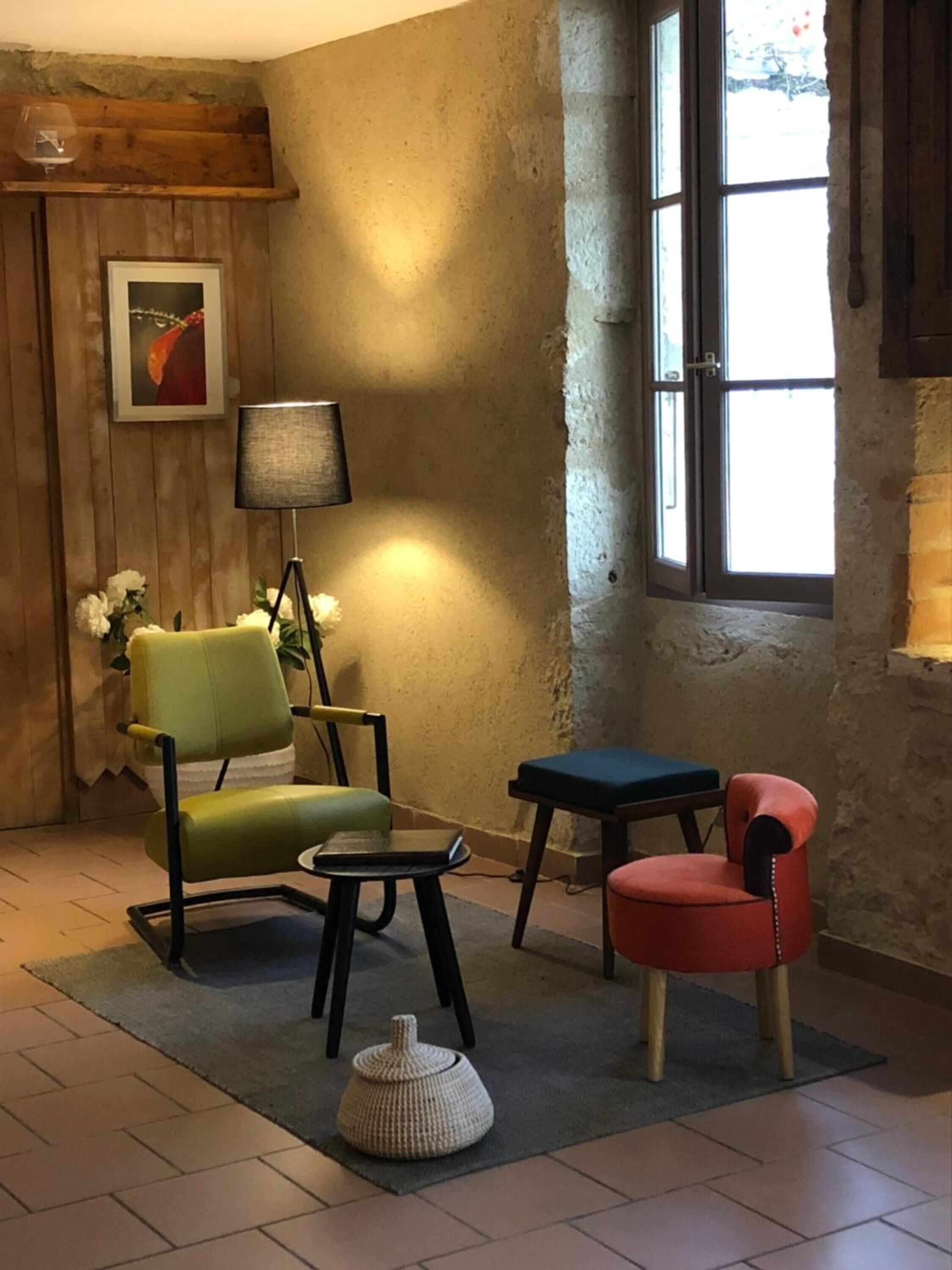 A cozy room with a wooden wall, a window, a green armchair, a red armchair, a small table, a lamp, and a vase with flowers.