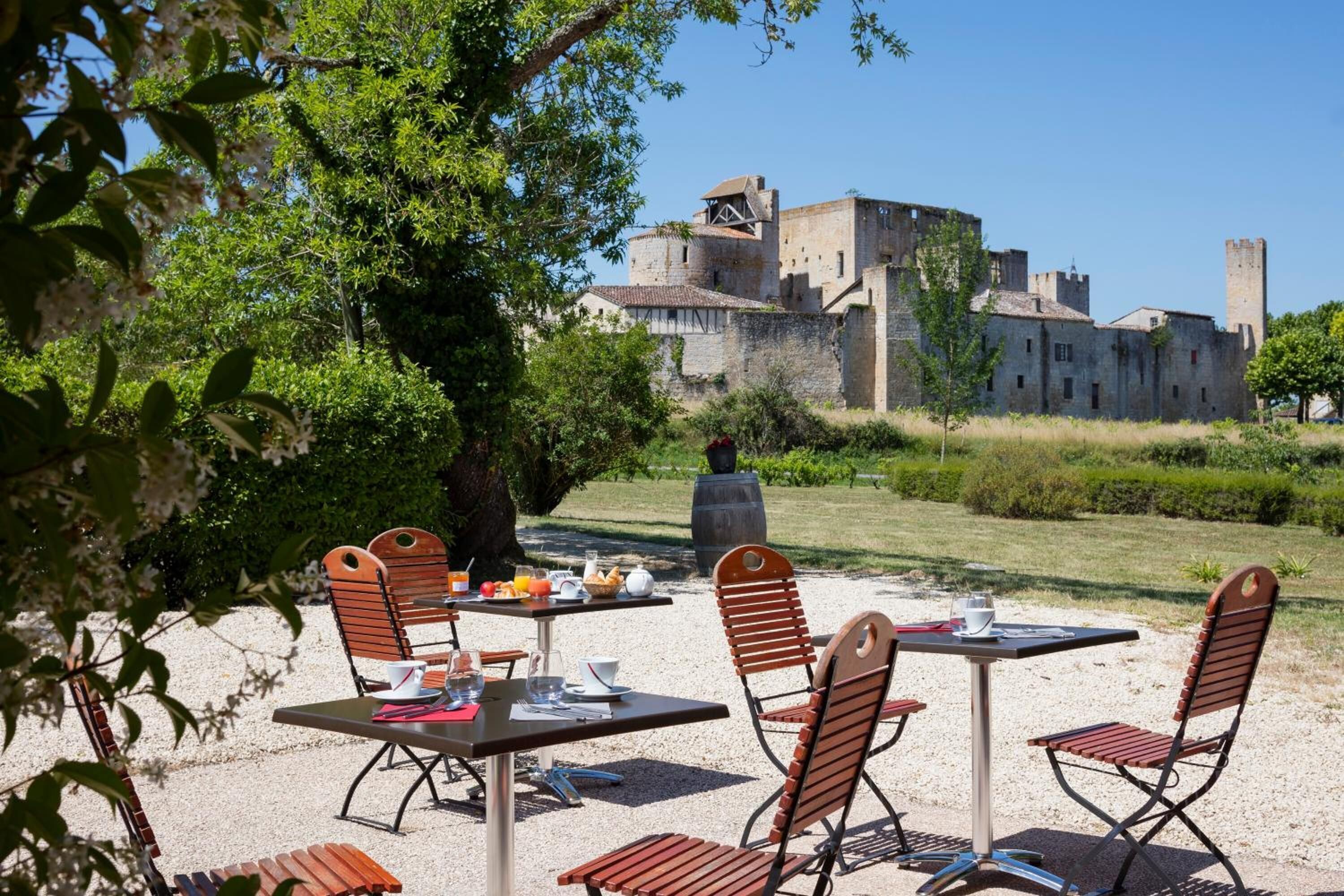 Un espace extérieur aménagé pour les repas, avec des tables et des chaises, offrant une vue sur un château historique en arrière-plan, et un arbre qui procure de l’ombre.