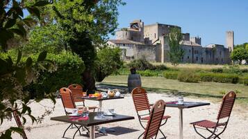 An outdoor dining area with tables and chairs, a view of a historic castle in the background, and a tree providing shade.