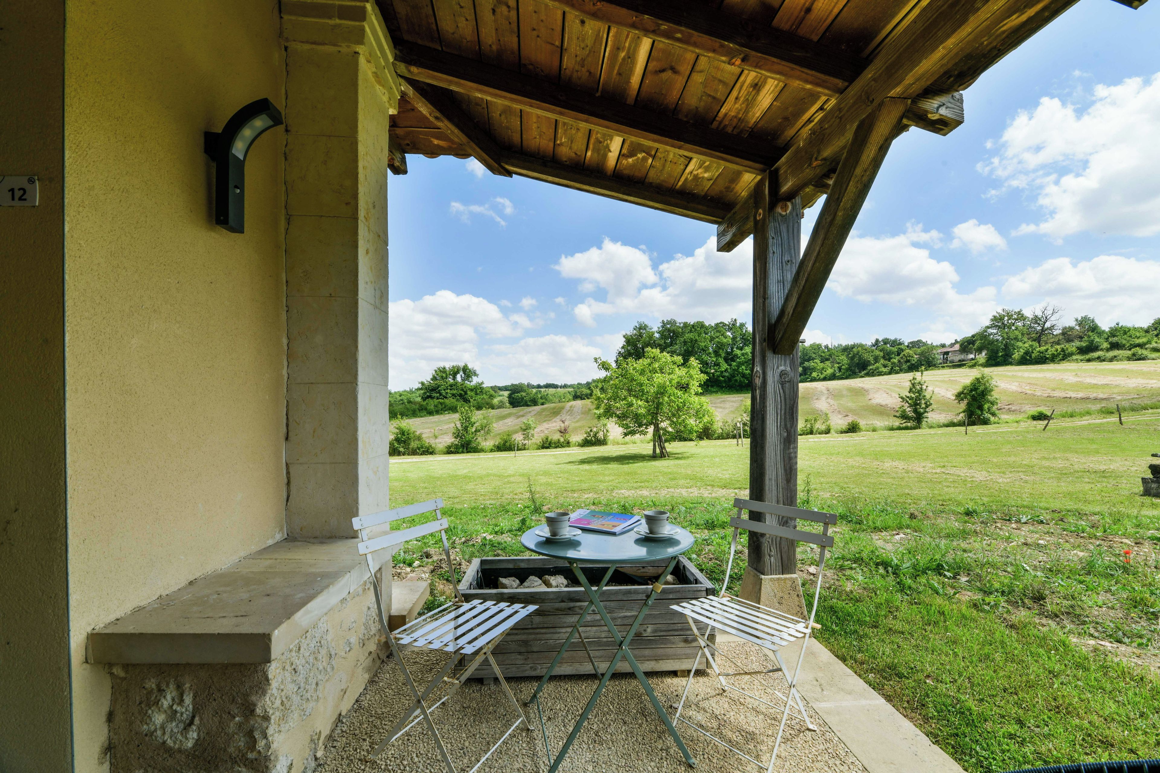 Een overdekt terras met een tafel en stoelen, uitzicht op een groen landschap en een gebouw met een nummerbord.