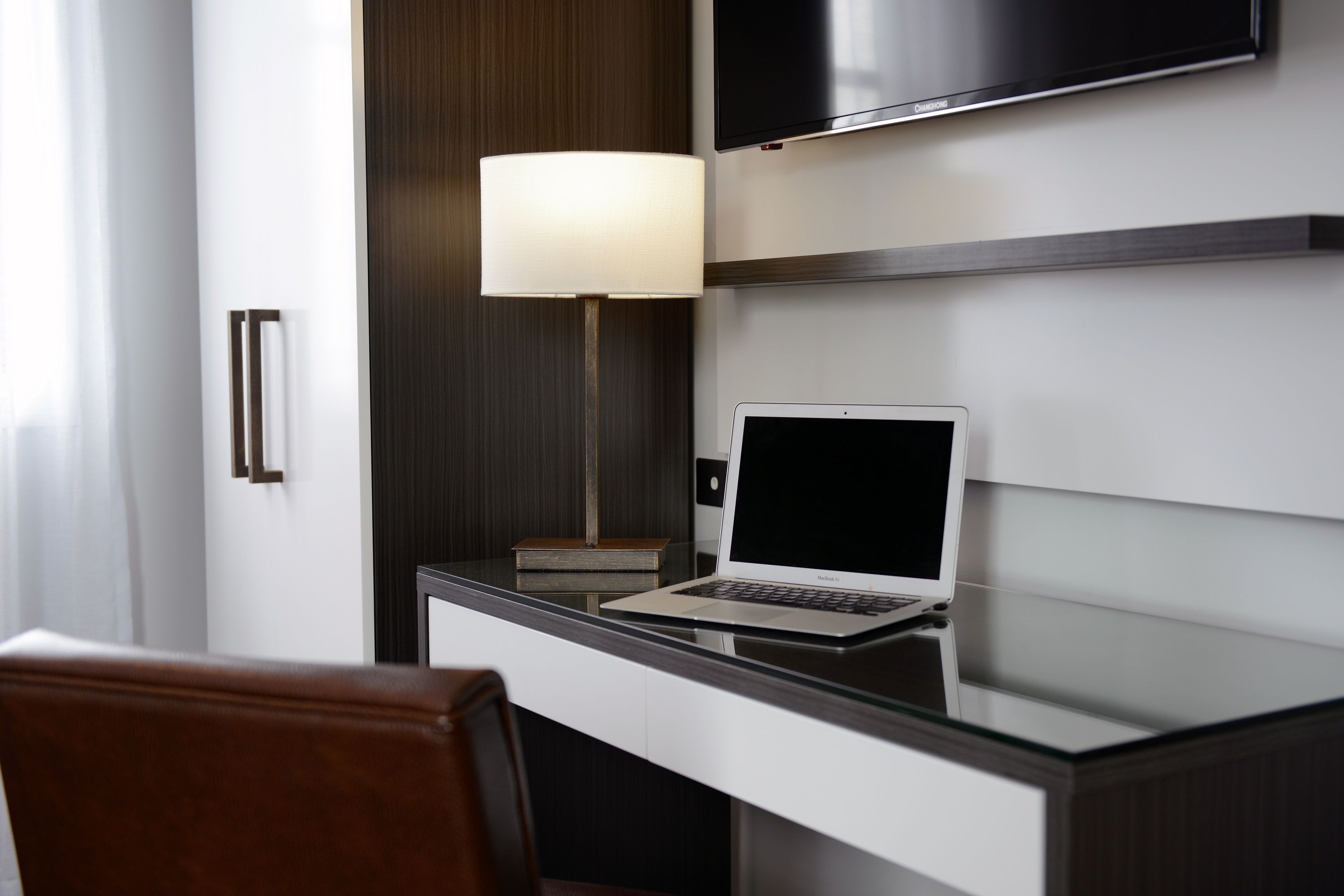 A modern hotel room with a laptop on a glass desk, a flat-screen TV mounted above, and a lamp on the desk.