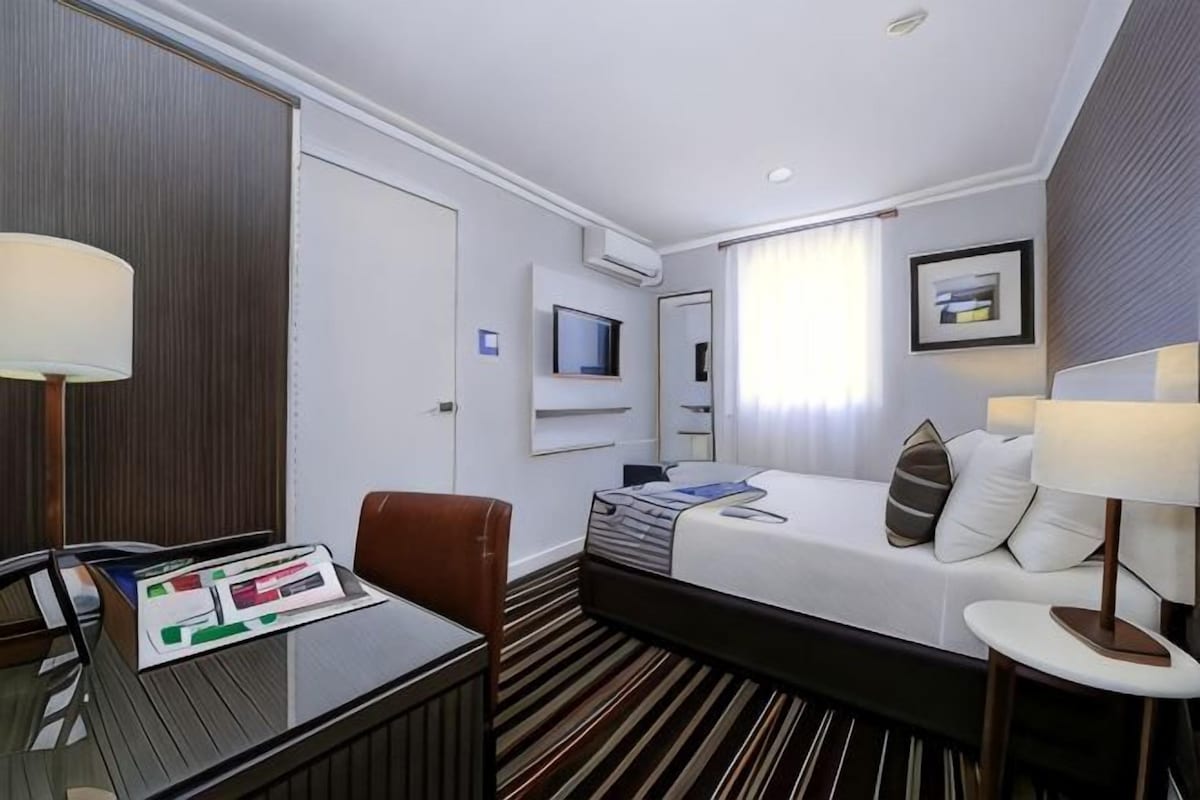 Habitación de hotel con cama, escritorio, silla, televisor y ventana con cortinas.