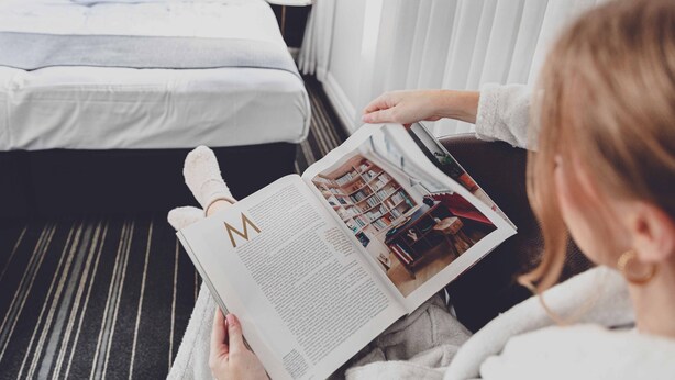 A person reading a magazine in a hotel room with a bed, a nightstand, and a window with blinds. - Perouse Randwick by Sydney Lodges (Randwick)