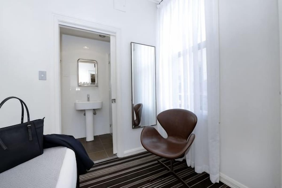 Habitación de hotel con cama, una silla, espejo, lavamanos y ventana con cortinas.