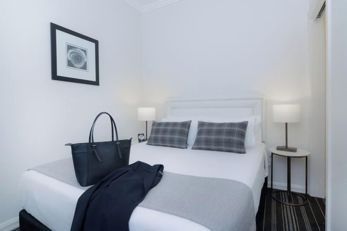 A hotel room with a neatly made bed, a black handbag, two bedside lamps, and a framed picture on the wall.