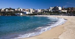 Una playa con aguas azules cristalinas, una orilla arenosa y un paisaje urbano al fondo.