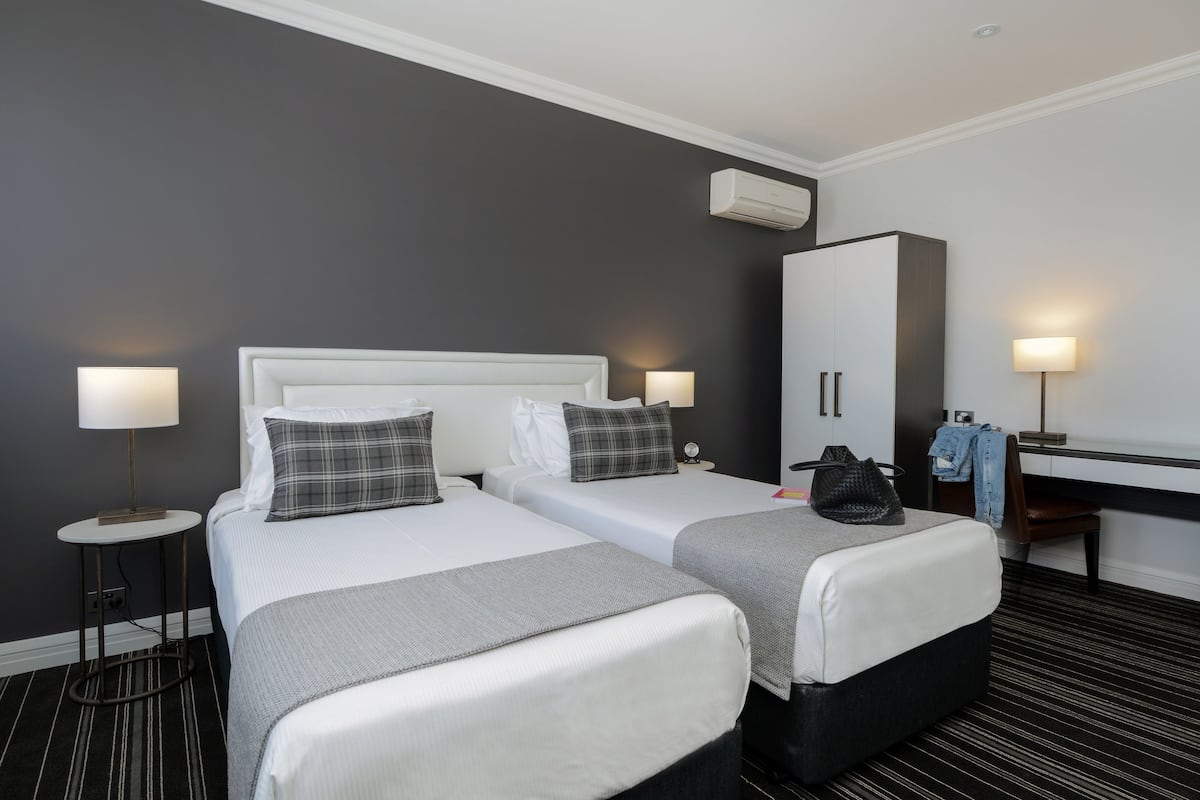 Una habitación de hotel con dos camas, un escritorio, un armario y una mesita de noche con una lámpara.