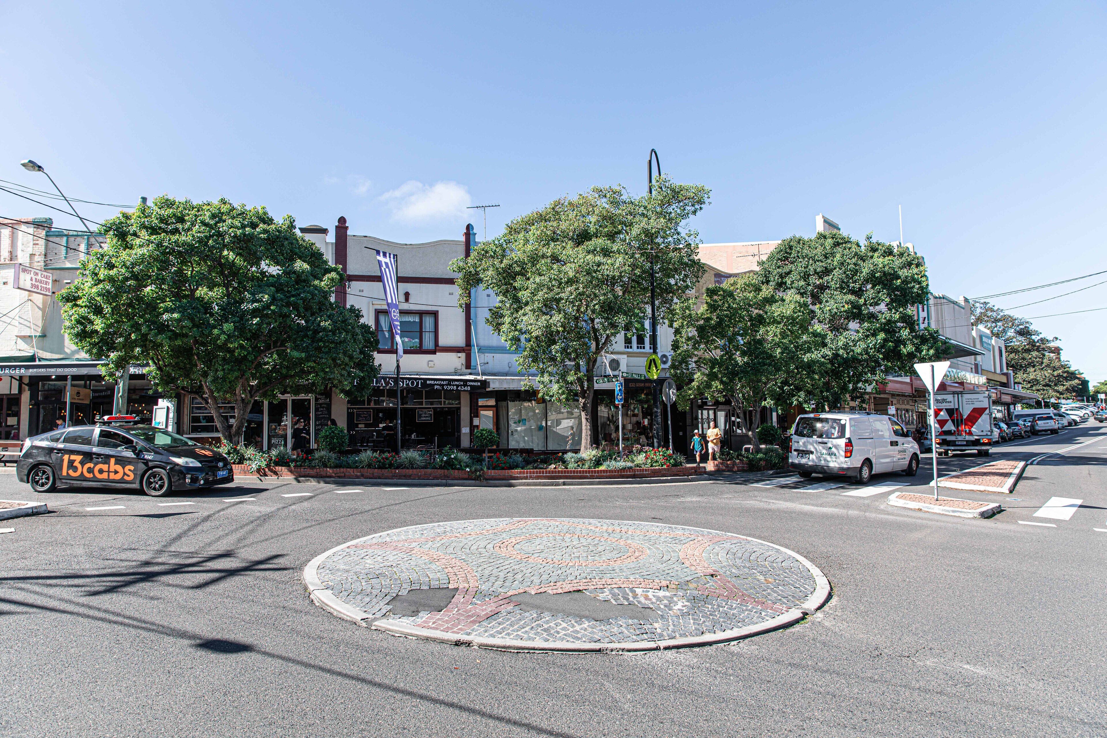 A roundabout with a cobblestone design, surrounded by trees and buildings with storefronts.