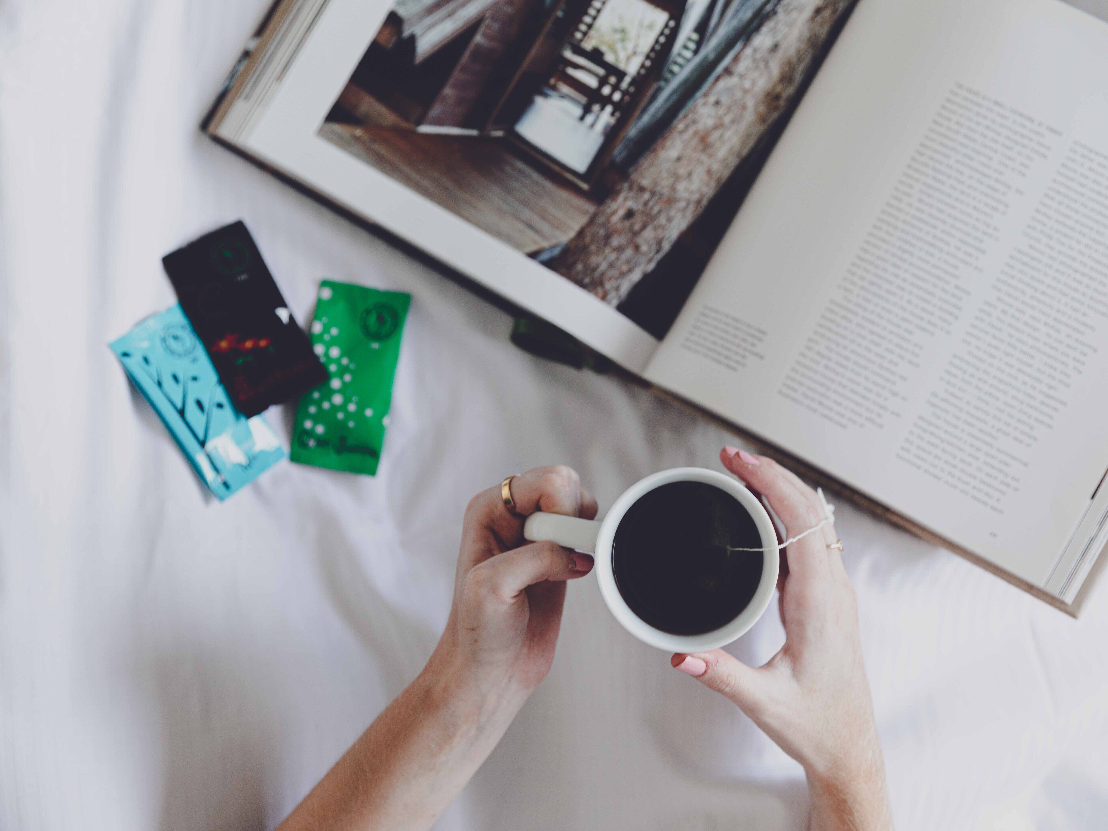 Uma pessoa segurando uma xícara de café, um livro e um smartphone em uma cama.