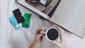 Uma pessoa segurando uma xícara de café, um livro e um smartphone em uma cama.