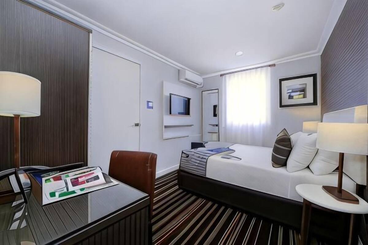 Habitación de hotel con cama, escritorio, silla, televisor y ventana con cortinas.