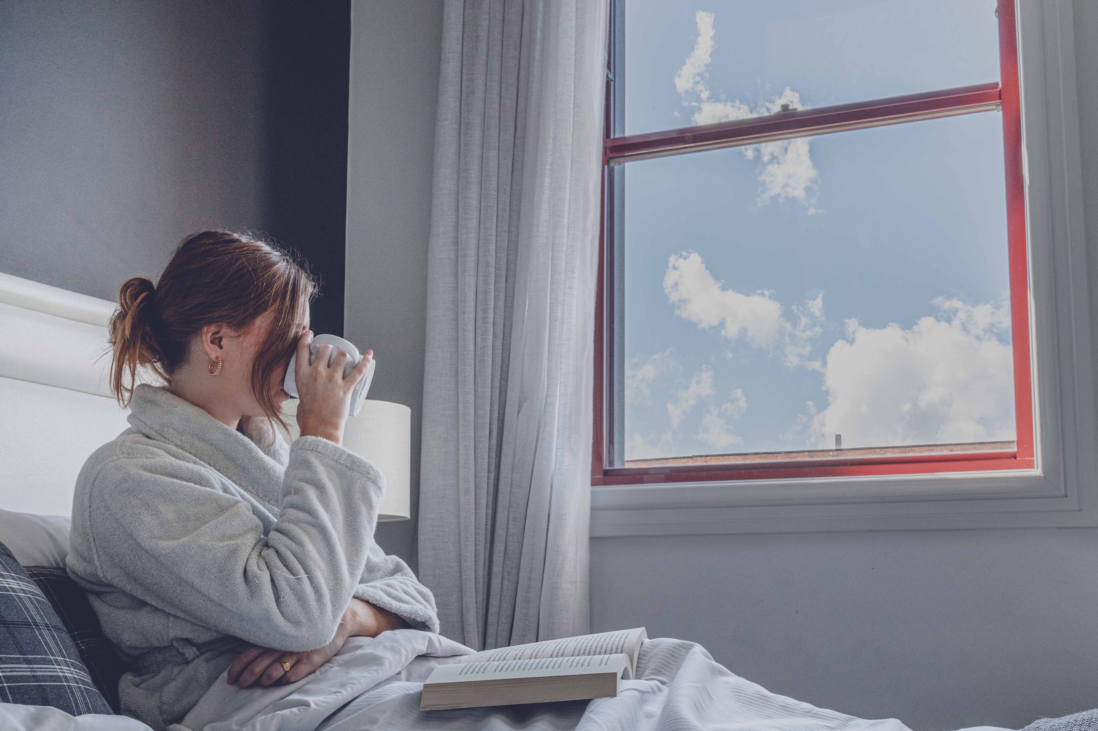 A person sitting on a bed with a book, a window with a view of the sky, and curtains.