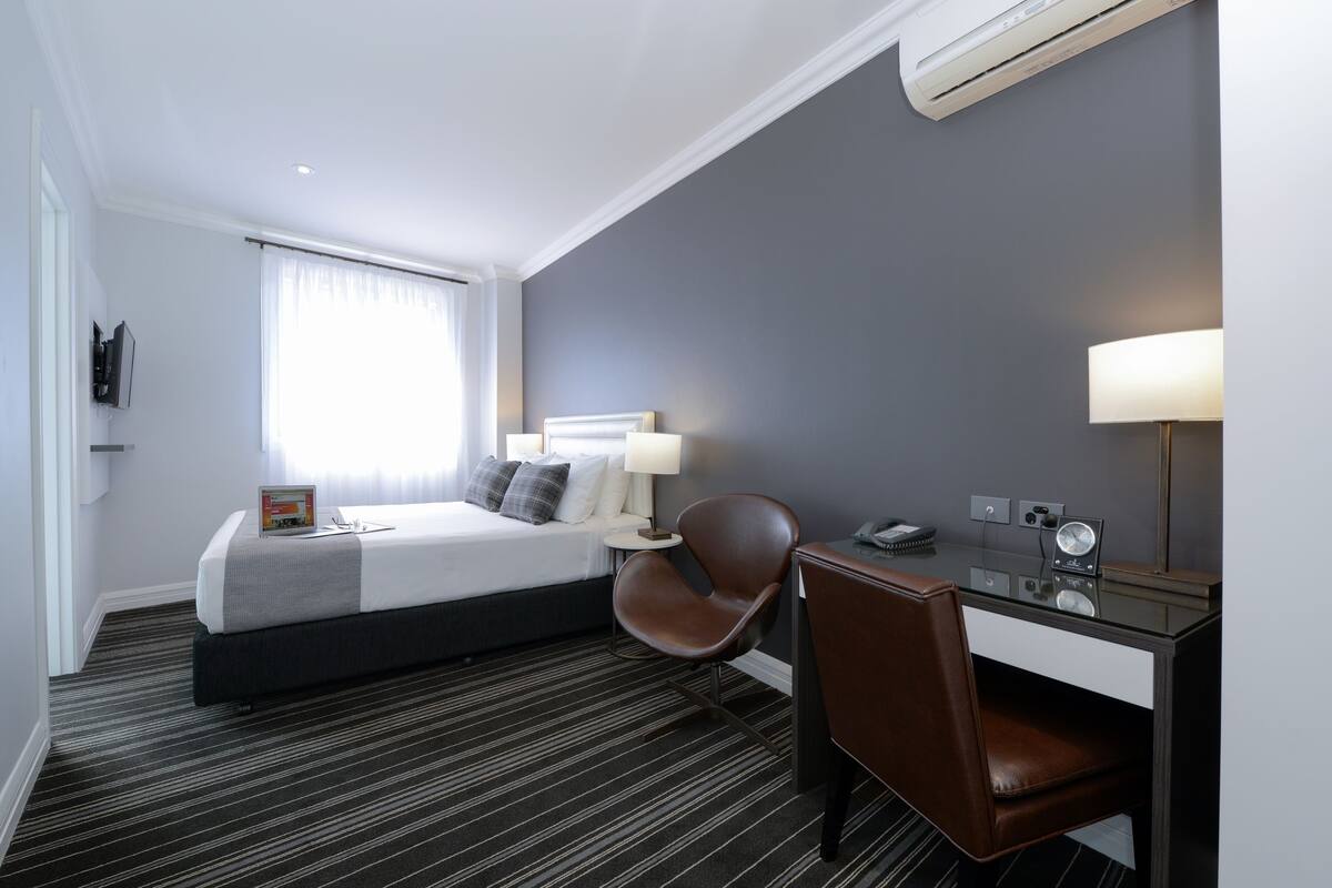 Una habitación de hotel con cama, escritorio, silla, lámpara y reloj.