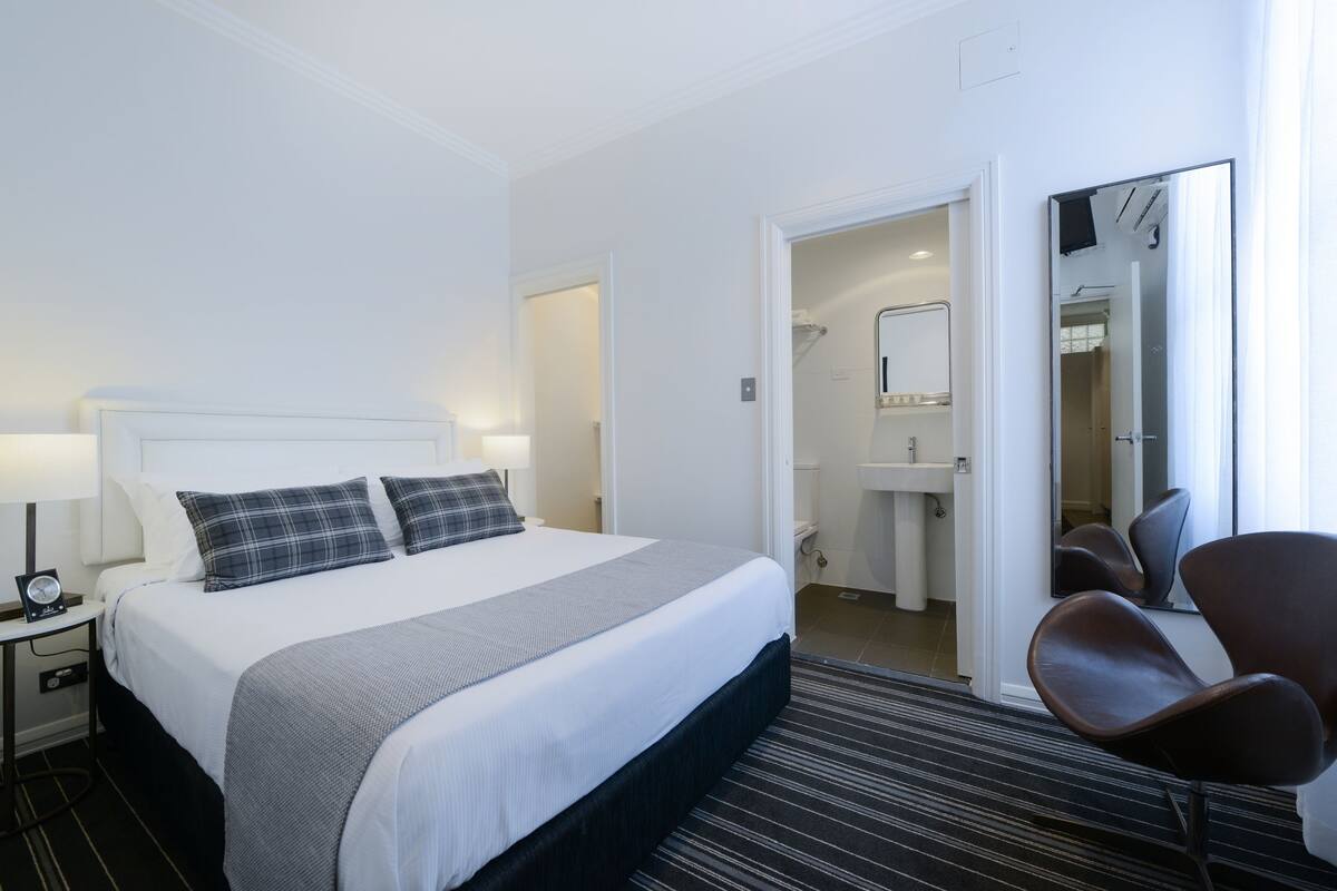 Habitación de hotel con una cama, una silla, una mesita de noche con lámpara, un baño con lavamanos y un espejo.