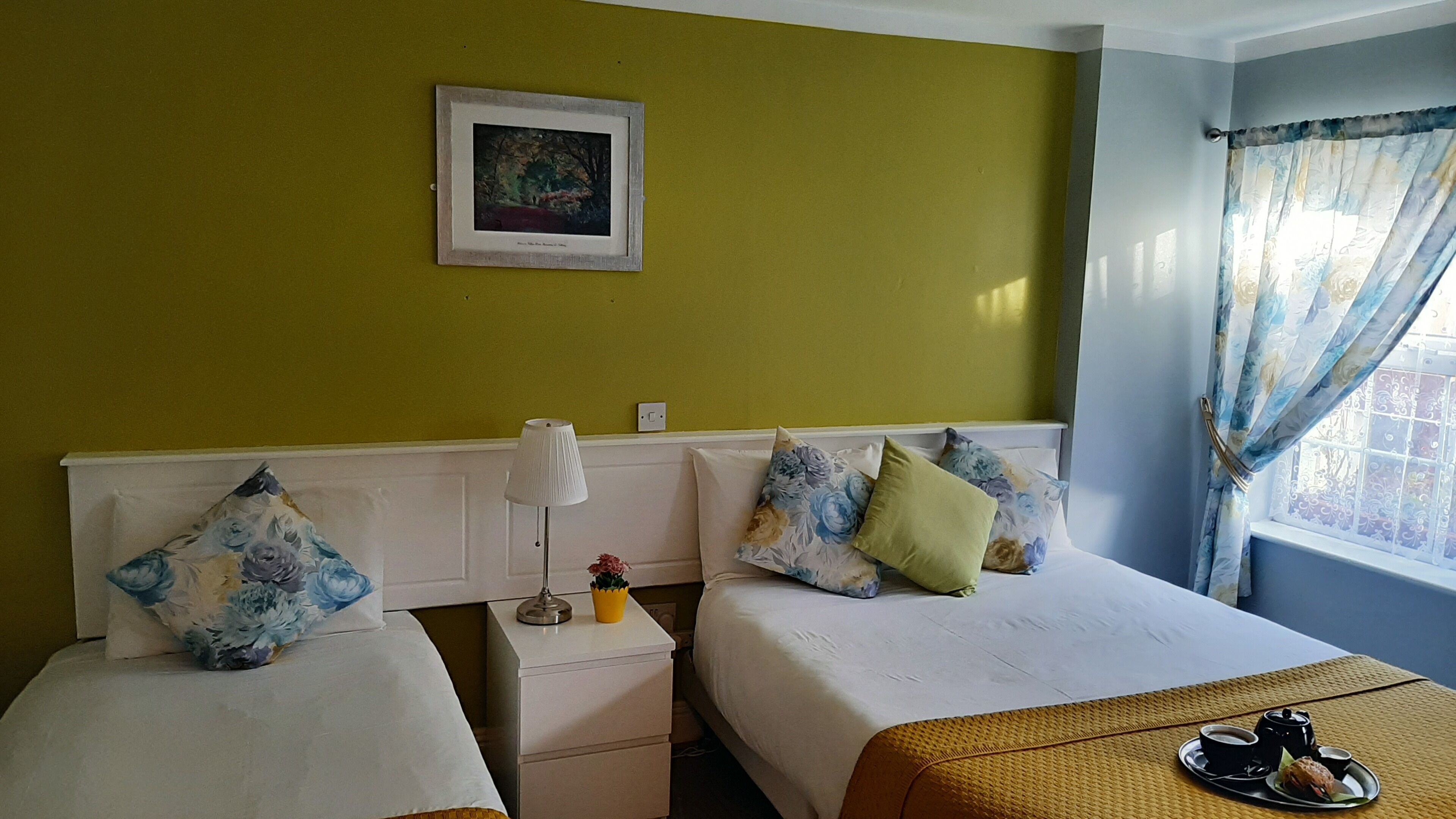 Habitación de hotel con dos camas, una mesita de noche con lámpara, un cuadro en la pared y una ventana con cortinas.