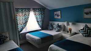 A hotel room with two beds, a window with chevron-patterned curtains, a bedside table with a lamp, and a wall-mounted artwork.