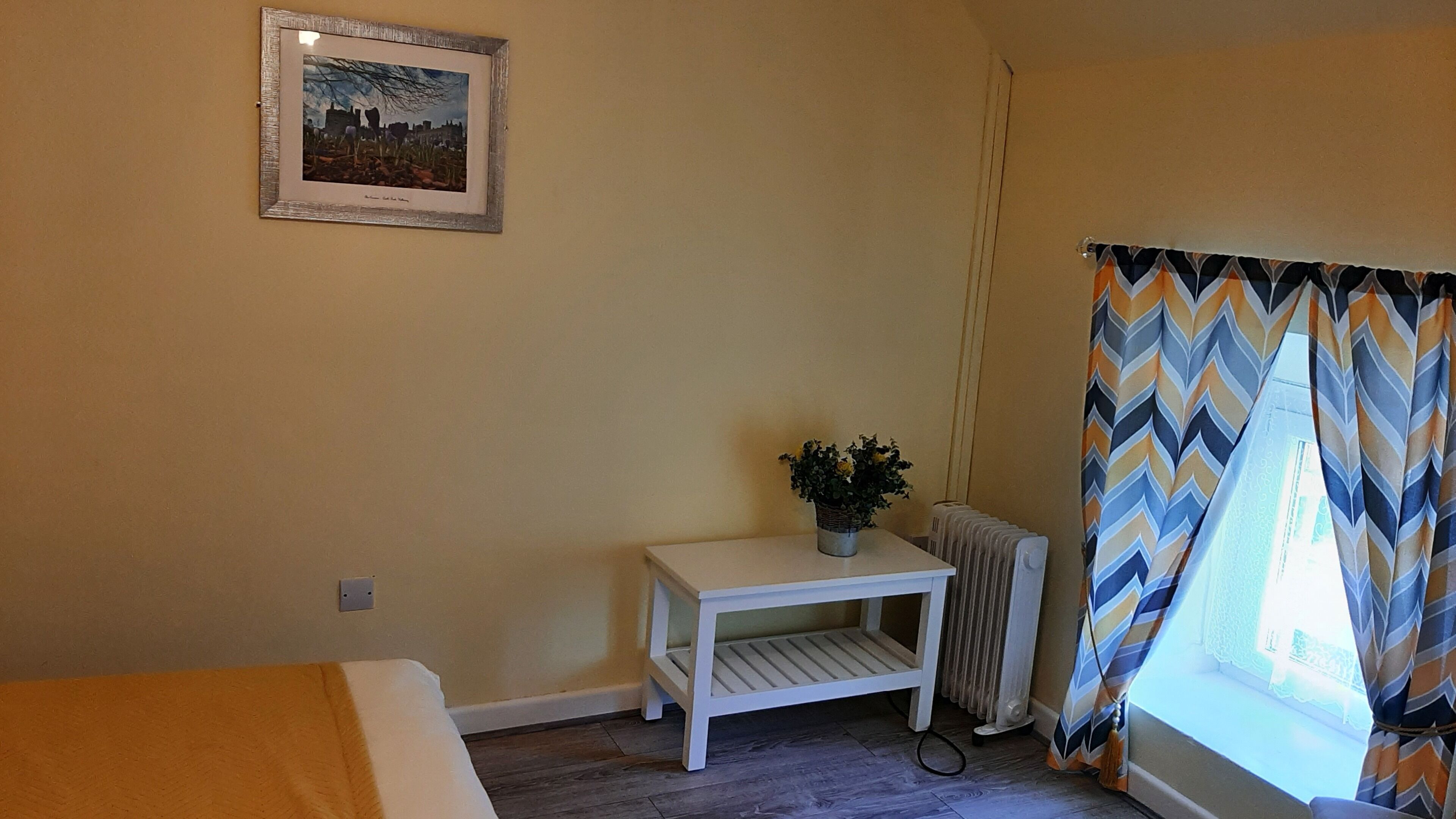 A small, cozy room with a bed, a small table with a plant, a radiator, and a window with patterned curtains.