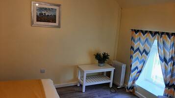A small, cozy room with a bed, a small table with a plant, a radiator, and a window with patterned curtains.