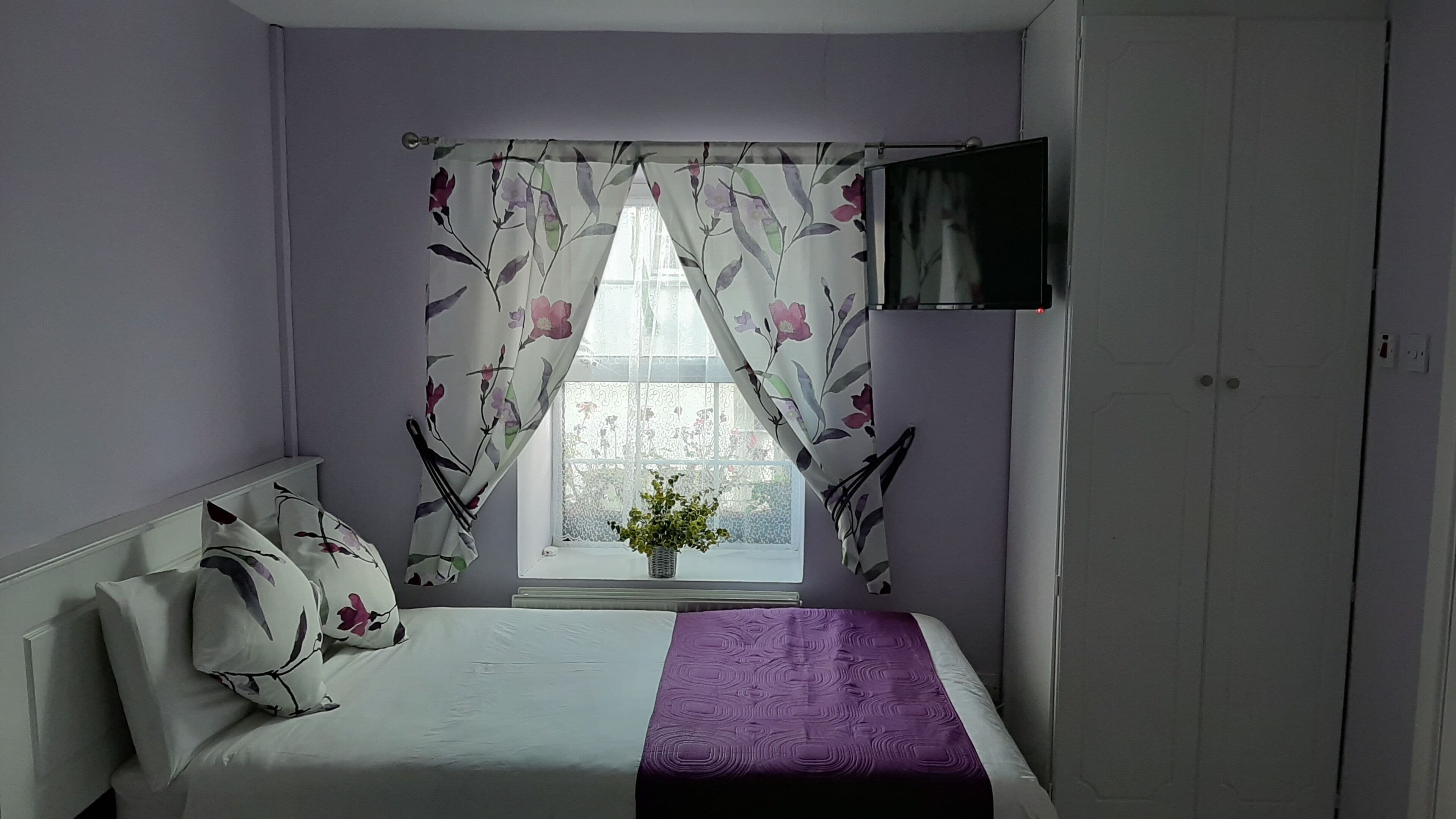 A bedroom with a bed, a window with floral curtains, a potted plant on the windowsill, and a TV mounted on the wall.