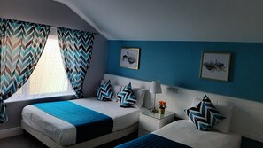 A hotel room with two beds, a window with chevron-patterned curtains, a nightstand with a lamp, and two framed floral artworks on the wall.