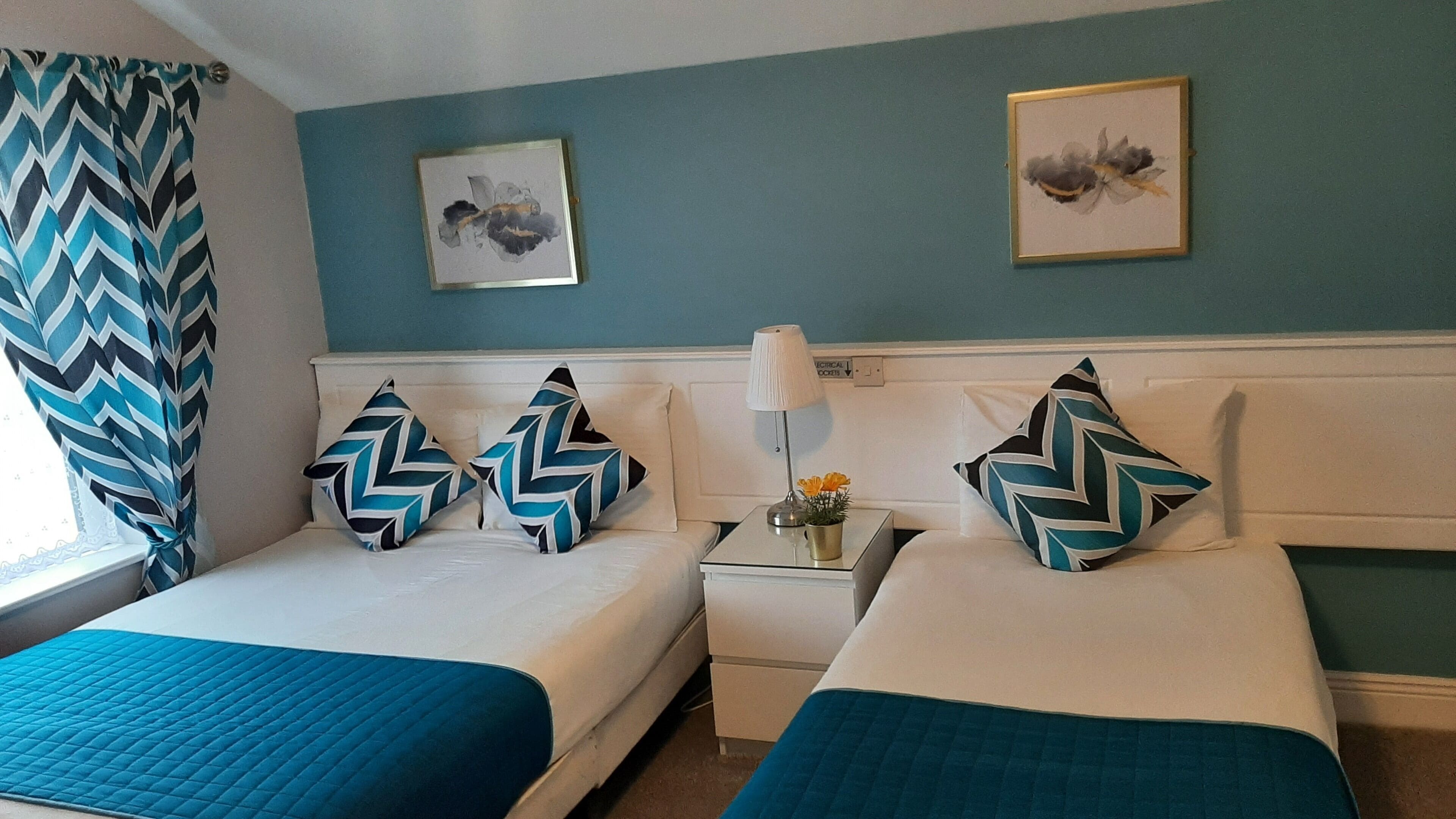 A hotel room with two beds, each with a teal quilted bedspread and matching pillows. There are two wall-mounted paintings, a nightstand with a lamp and a vase of flowers, and a window with a chevron-patterned curtain.