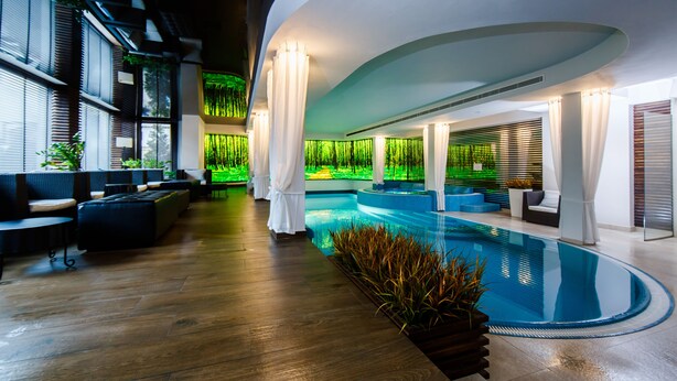 A modern indoor swimming pool with wooden flooring, seating areas, and green wall decor.