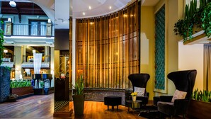 A lobby with wooden flooring, a seating area with black chairs, a large wooden wall, and a balcony with plants.