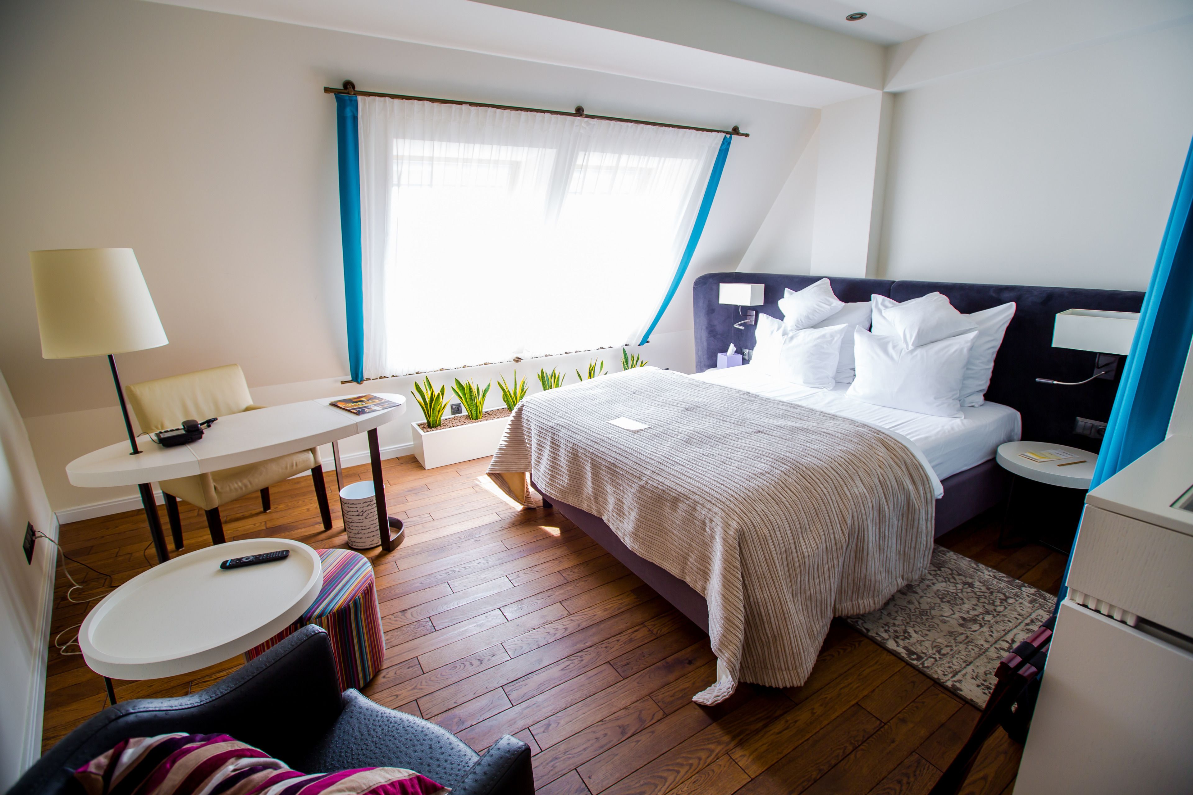 A hotel room with a bed, a desk, a chair, a small table, and a window with curtains.