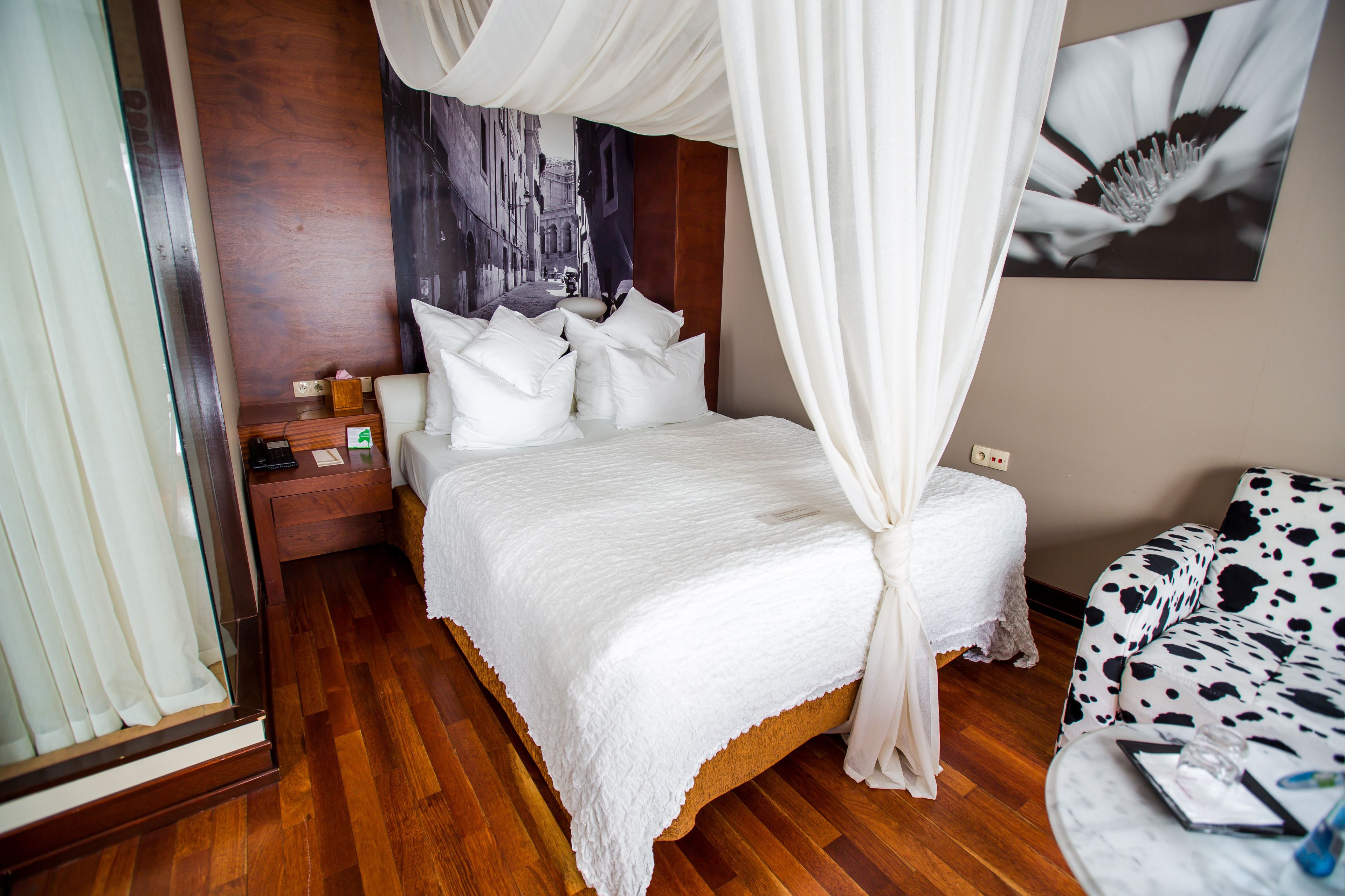 A bedroom with a canopy bed, wooden flooring, a nightstand, a bedside table, a patterned sofa, and a wall-mounted picture.