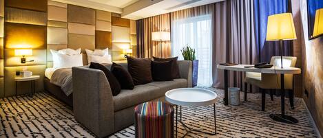Una habitación de hotel con una cama, un sofá, una mesita redonda pequeña, una silla y una mesita de noche con una lámpara.
