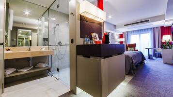 A modern hotel room with a glass shower, a vanity with a mirror, a bed with a dark headboard, and a red accent wall.