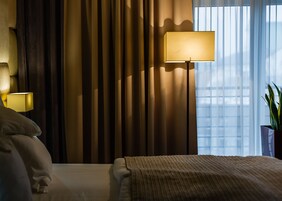 Una habitación de hotel con cama, lámpara de noche, cortinas y ventana con persianas.