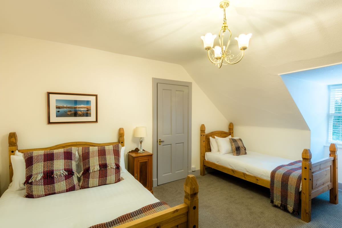 A bedroom with two single beds, a wooden headboard, a small bedside table with a lamp, a framed picture on the wall, and a window with blinds.