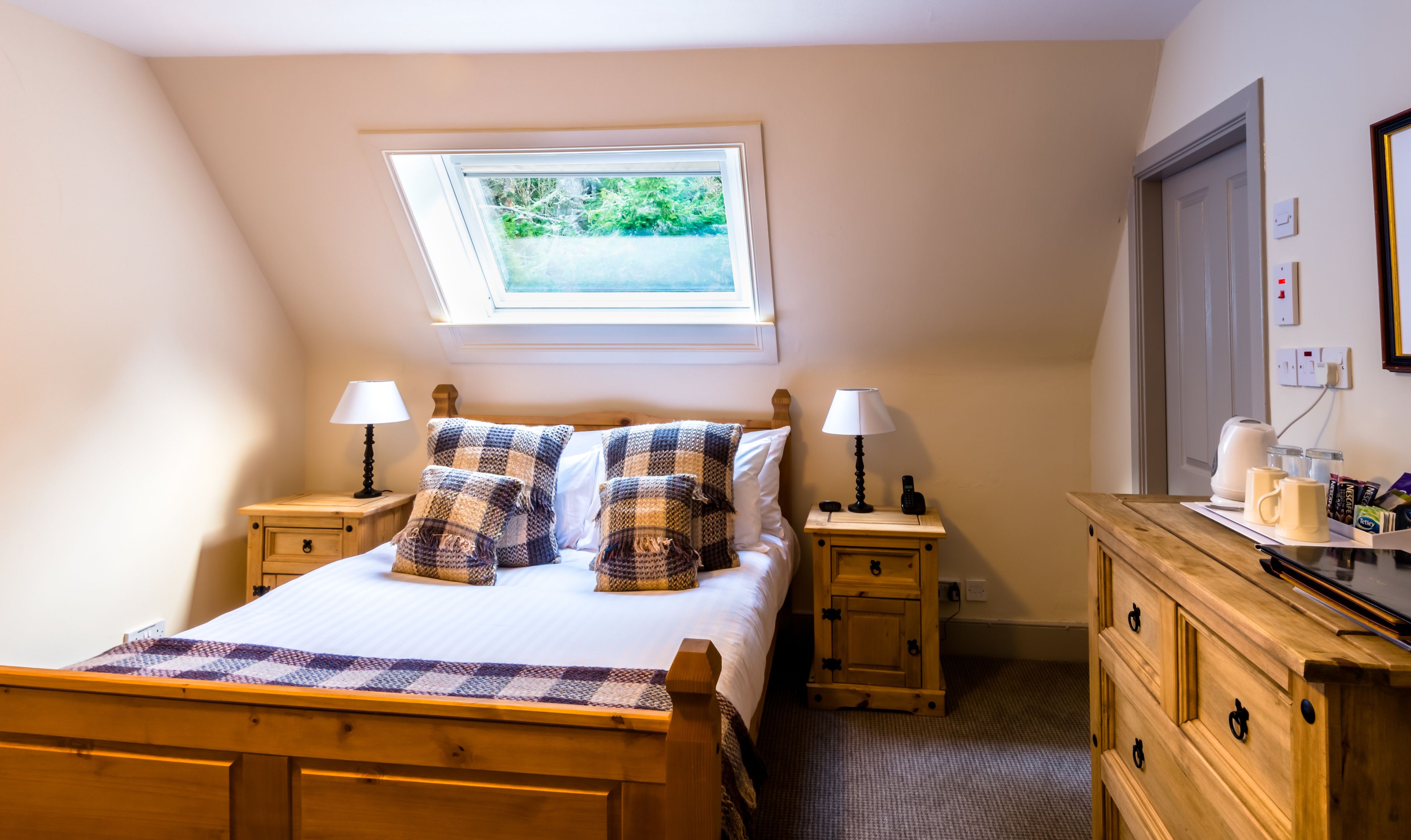 A bedroom with a bed, two nightstands, a wooden dresser, a skylight, and a view of greenery outside.