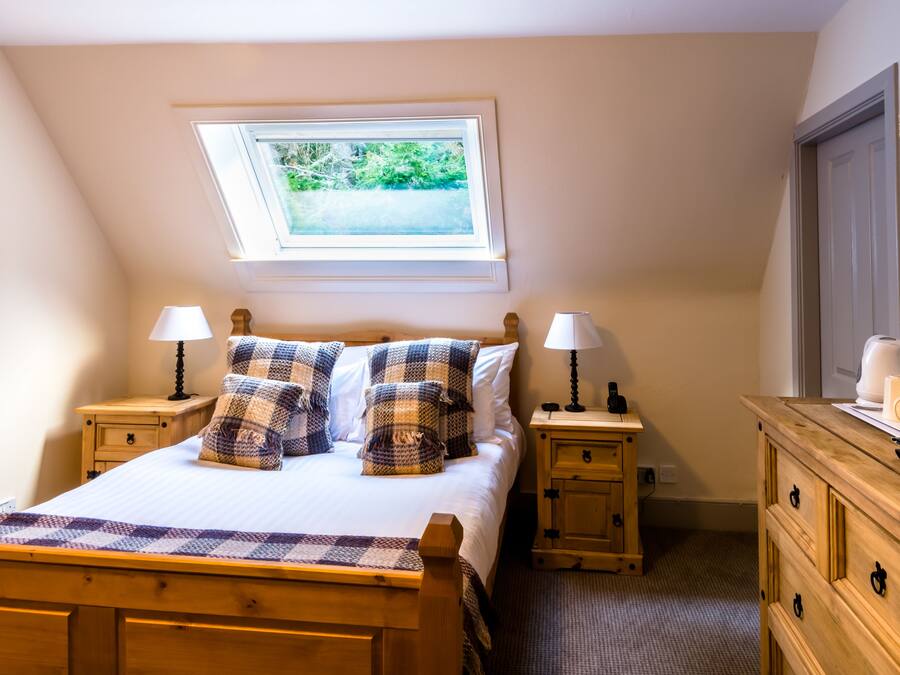 A bedroom with a bed, two nightstands, a wooden dresser, a skylight, and a view of greenery outside.