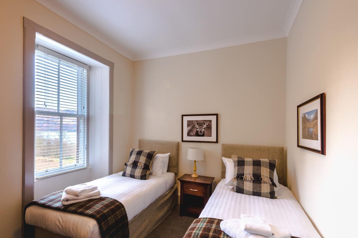 A hotel room with two beds, a window with blinds, a nightstand with a lamp, and framed pictures on the wall.