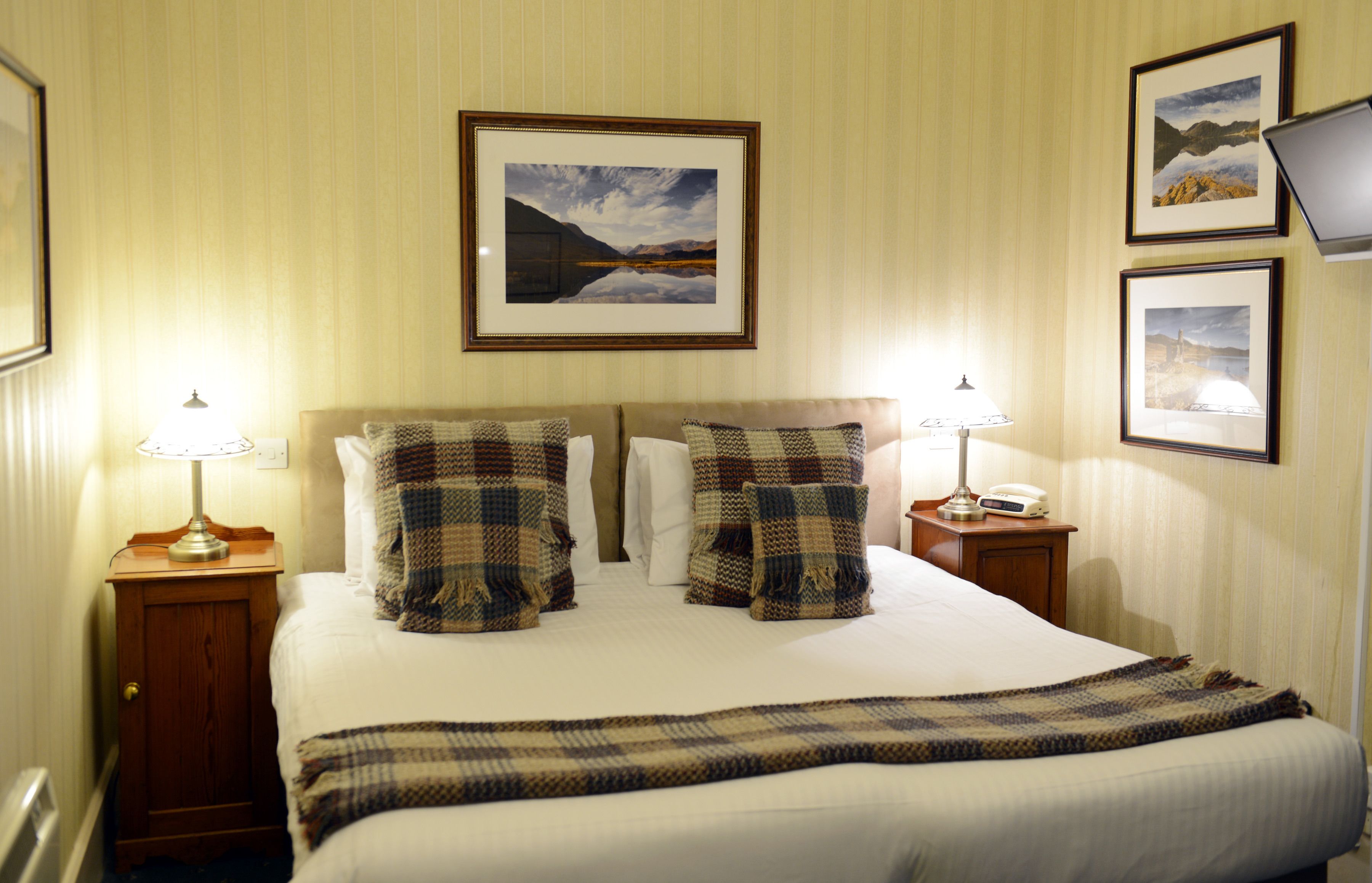 A hotel room with a large bed, two bedside tables with lamps, a framed landscape picture above the bed, and a second framed picture on the wall.