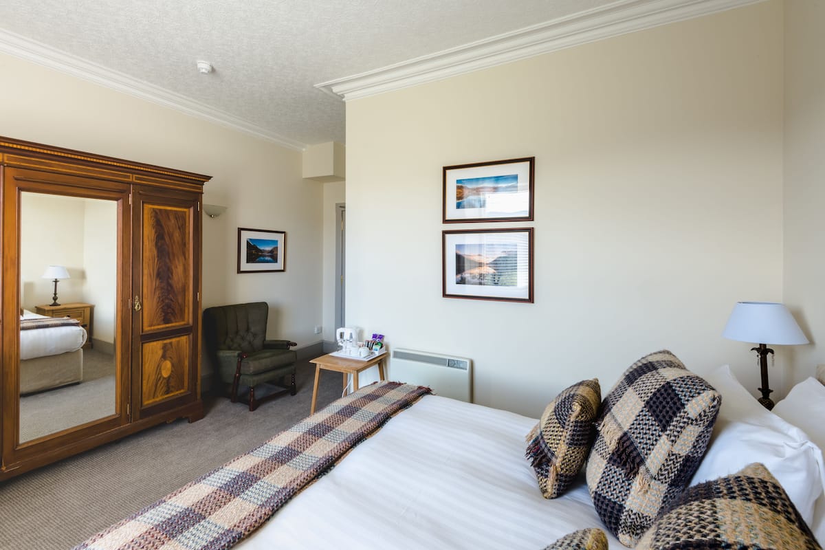 A hotel room with a large bed, a wardrobe, a chair, a small table with a lamp, and two framed pictures on the wall.
