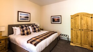 A neatly made bed with a checkered blanket, two framed pictures on the wall, a wooden wardrobe, and a small bedside table with a lamp. - The Inch Hotel (Fort Augustus)