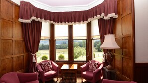 A room with two armchairs, a table, and a lamp. There are large windows with curtains, and a view of the outdoors. - The Inch Hotel (Fort Augustus)