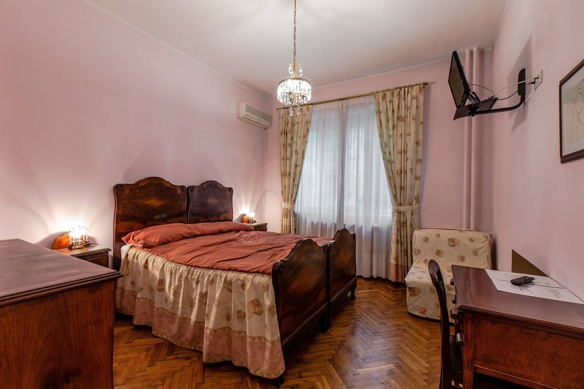 A bedroom with a wooden bed, a desk, a chair, a chandelier, and a window with curtains.