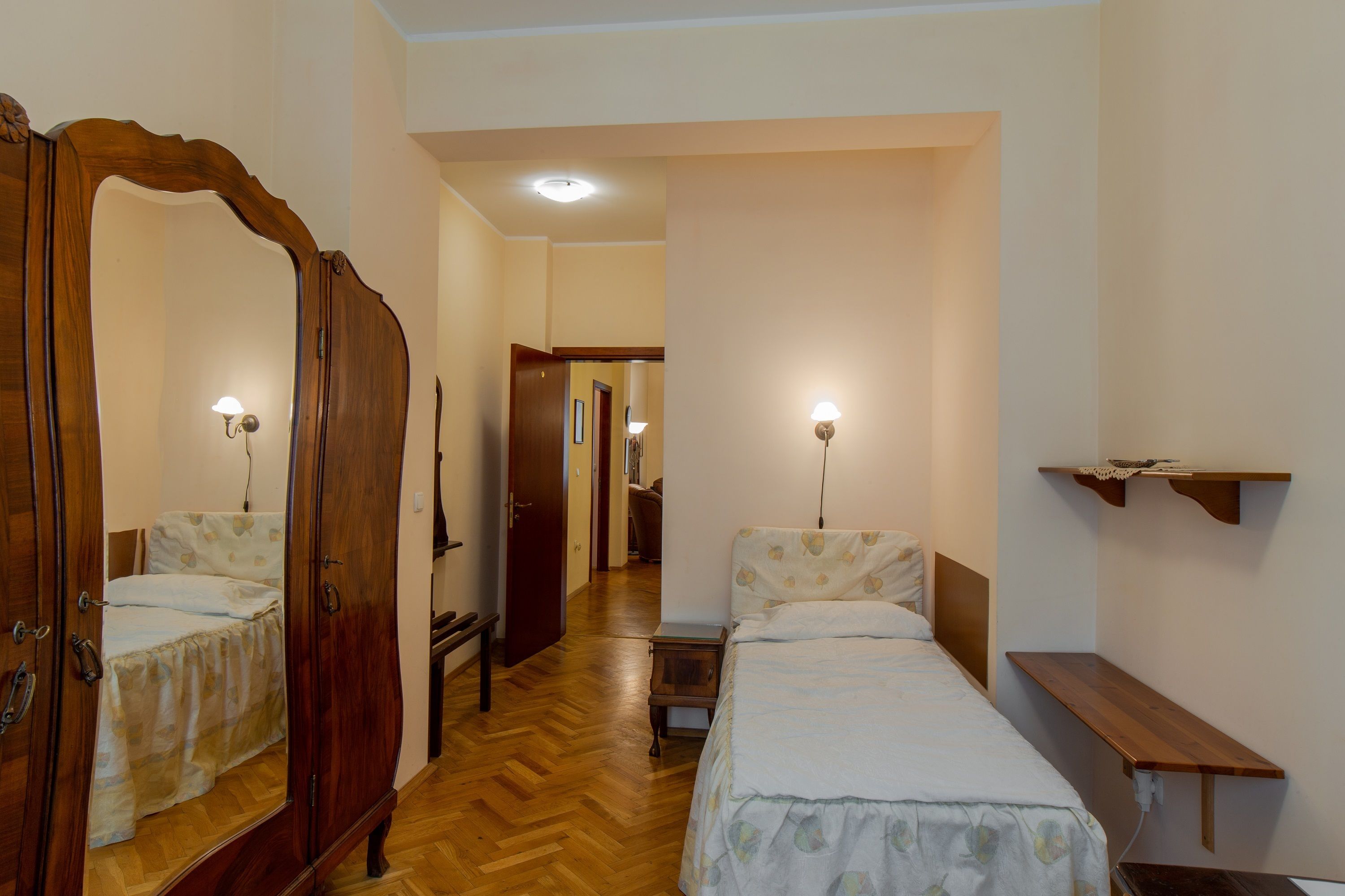 A bedroom with a wooden wardrobe, a bedside table, a bed with a floral bedspread, a nightstand, and a wall-mounted shelf.