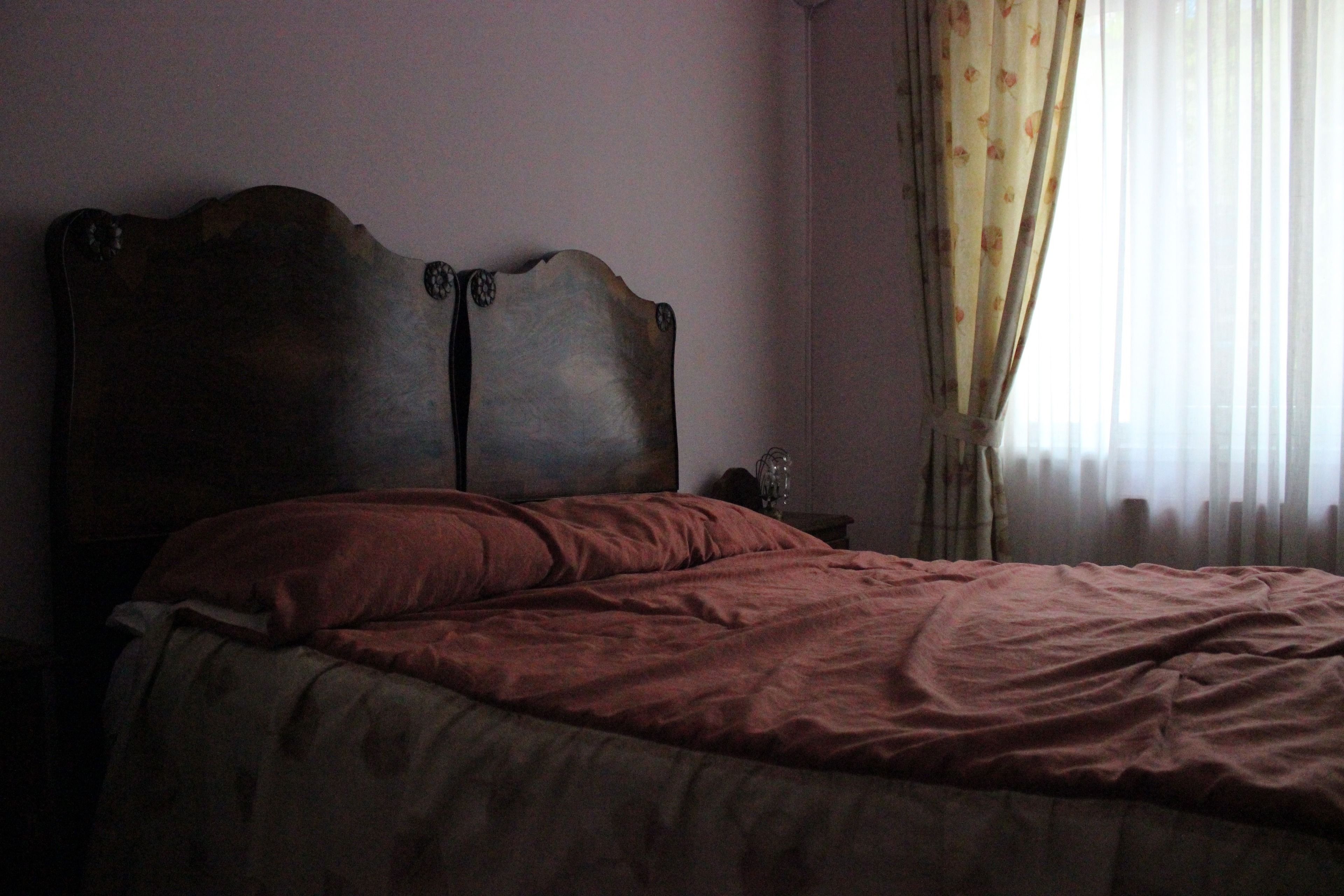 Un dormitorio con cabecera de madera, cama con manta roja, mesita de noche con lámpara y ventana con cortinas.