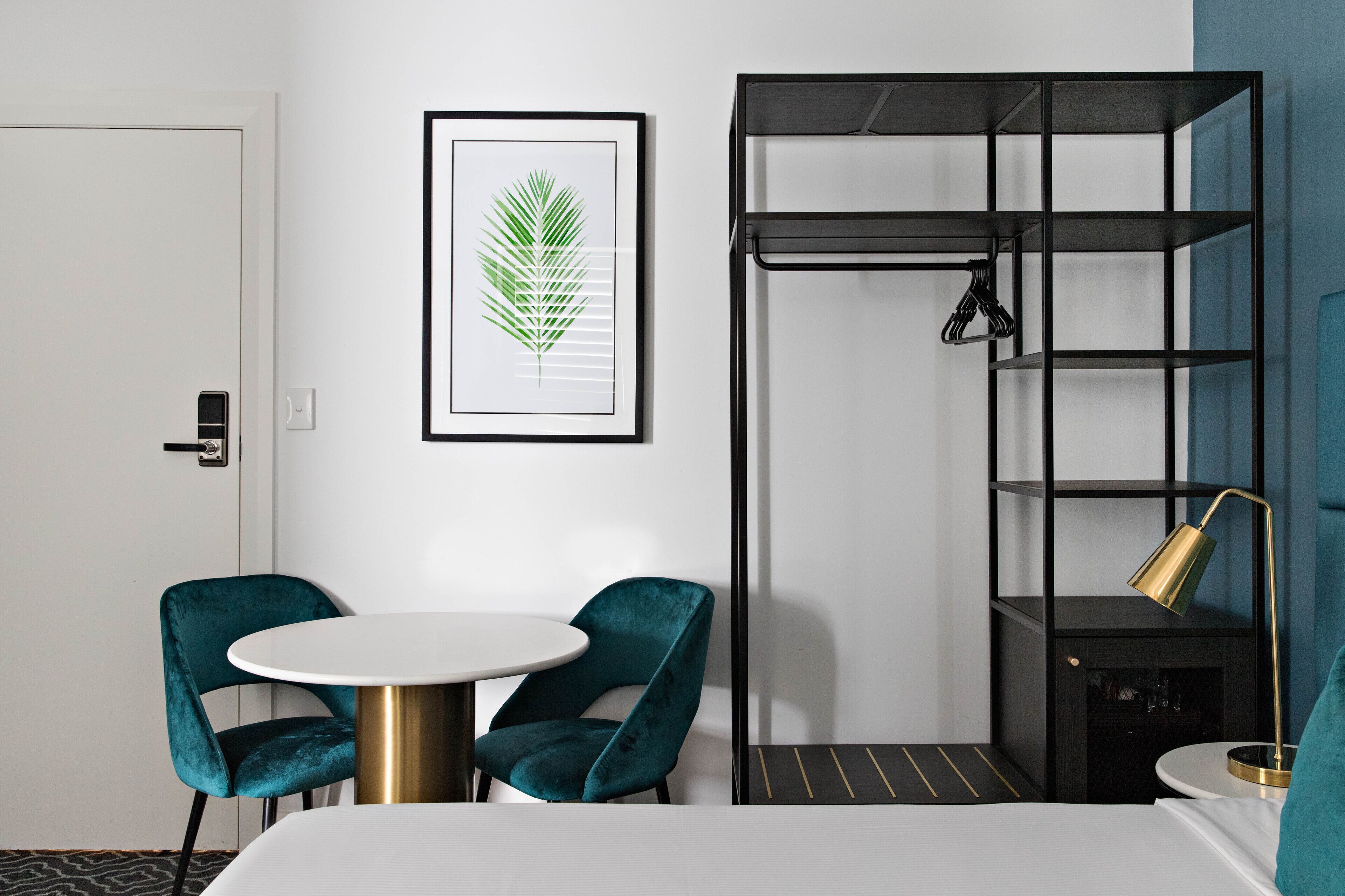 Ein Hotelzimmer mit einem Bett, zwei smaragdgrünen Sesseln, einem runden Tisch, einer gerahmten Blattform-Kunstwerk, einer schwarzen Regaleinheit und einer goldfarbenen Lampe.