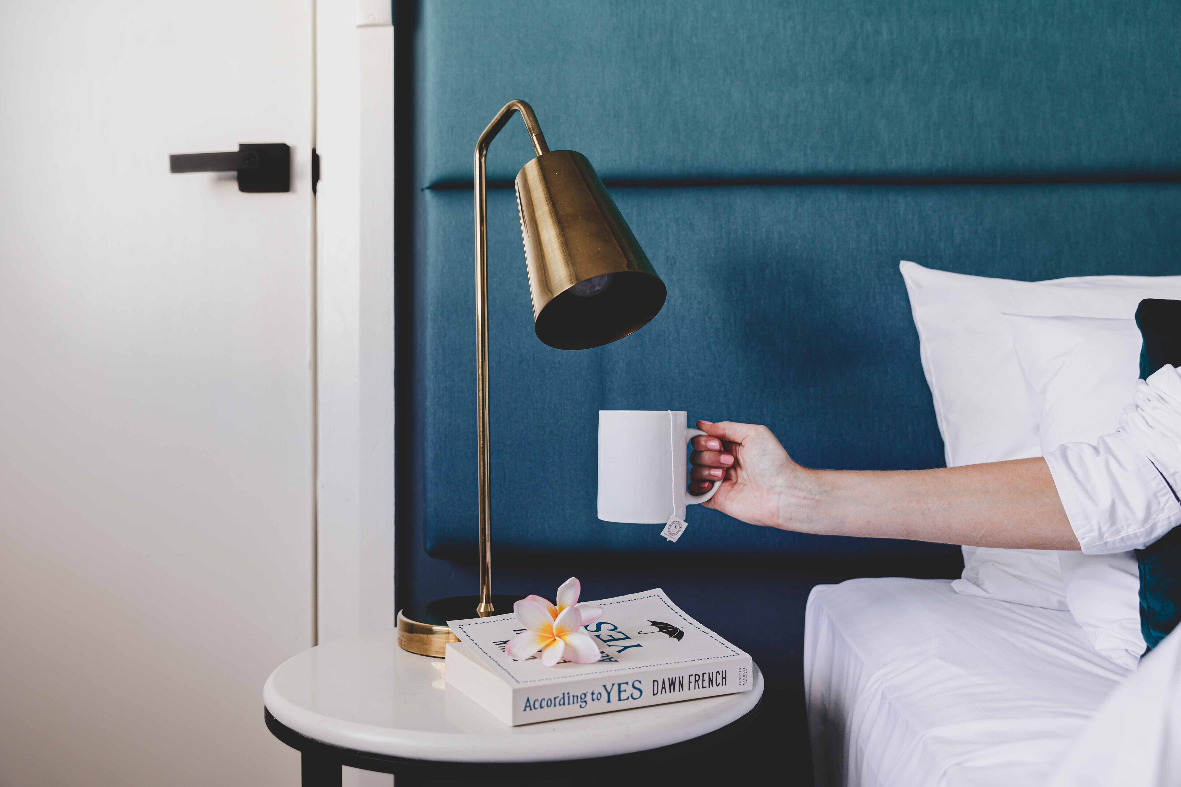 A neatly made bed with white bedding, a bedside table with a book and a flower, and a gold-colored lamp.