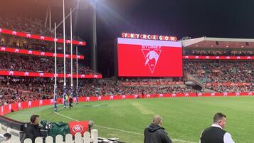 Un estadio con múltiples niveles de gradas, una gran pantalla digital que muestra el logotipo del Sydney Cricket Ground, y un campo verde.